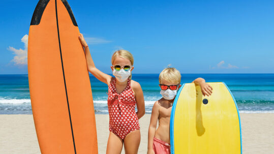 Children on beach with surf boards wearing COVID face masks